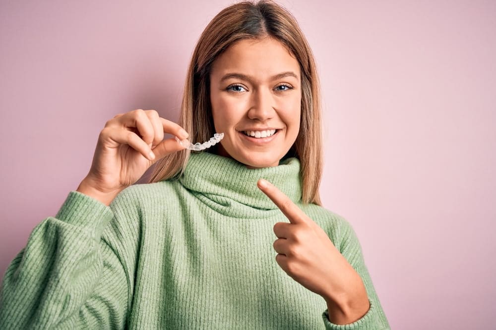 Smiling woman in green sweater holds Invisalign aligner, representing Carpinello Orthodontics in Drexel Hill, PA.
