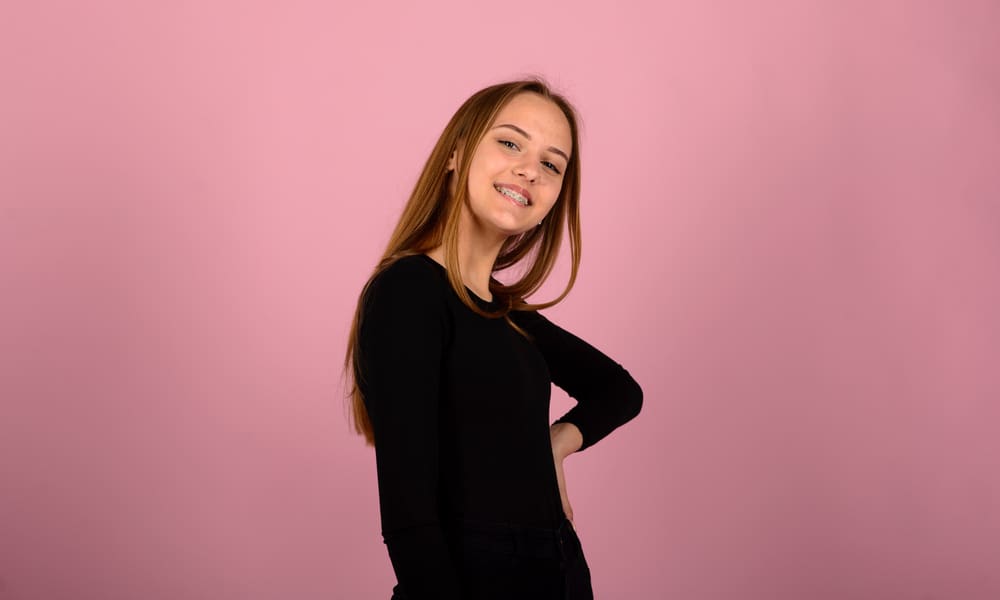 Smiling teen girl with braces poses in black shirt on pink backdrop for Carpinello Orthodontics in Edgmont, PA.