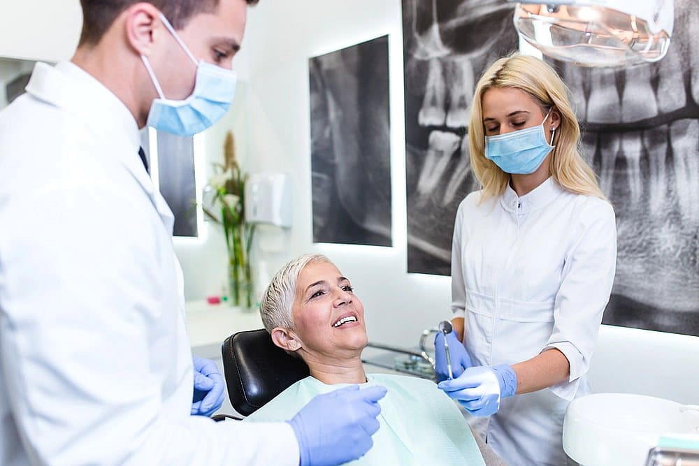 A smiling patient at Carpinello Orthodontics in Drexel Hill, PA, is treated by two masked dental professionals with orthodontic appliances.