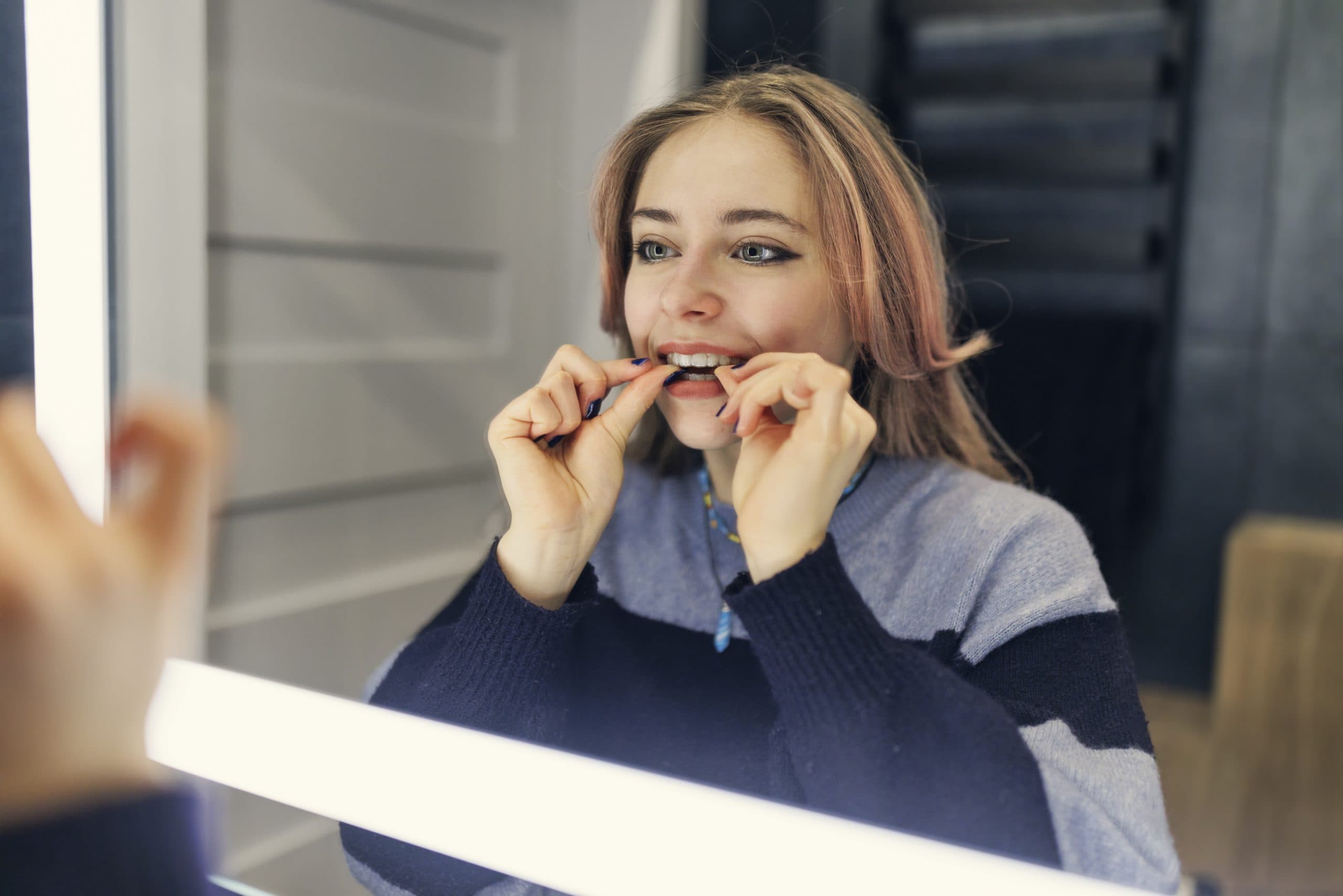 A woman flosses her teeth in front of a mirror, showing Invisalign aligners from Carpinello Orthodontics in Edgmont, PA.