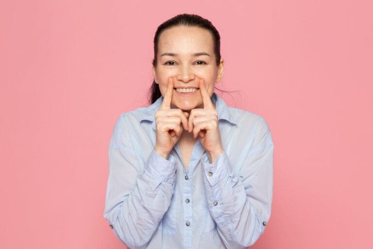 At Carpinello Orthodontics in Drexel Hill, Newtown Square or Edgmont, PA, woman in blue shirt playfully smiles against pink backdrop.