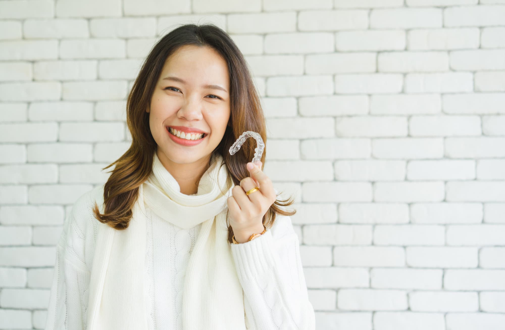 Smiling woman in white sweater and scarf holds Invisalign® Clear Aligners at Carpinello Orthodontics in Drexel Hill, PA.