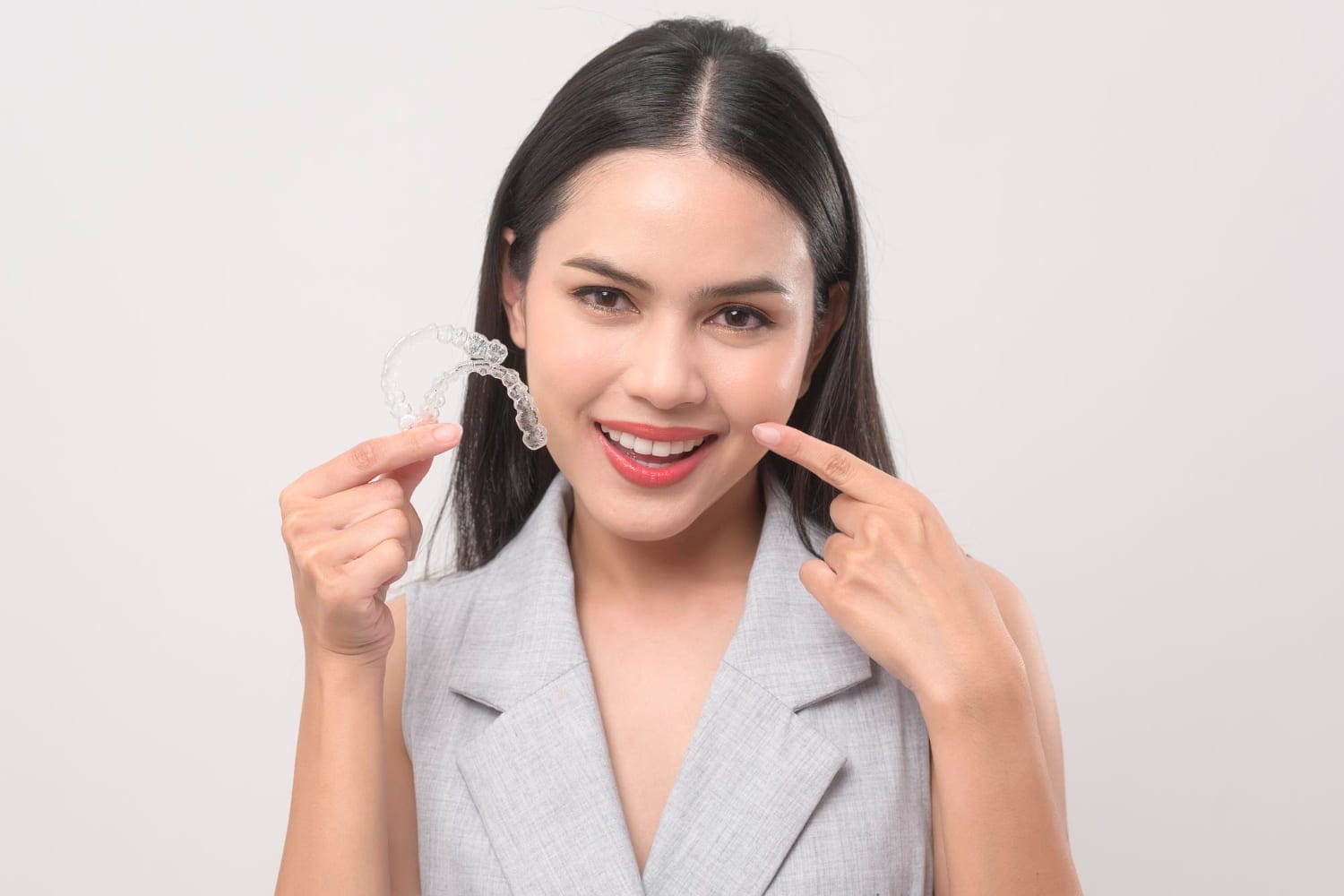 Smiling woman in a gray sleeveless blazer holds Invisalign® Clear Aligners, promoting Carpinello Orthodontics in Newtown Square, PA.