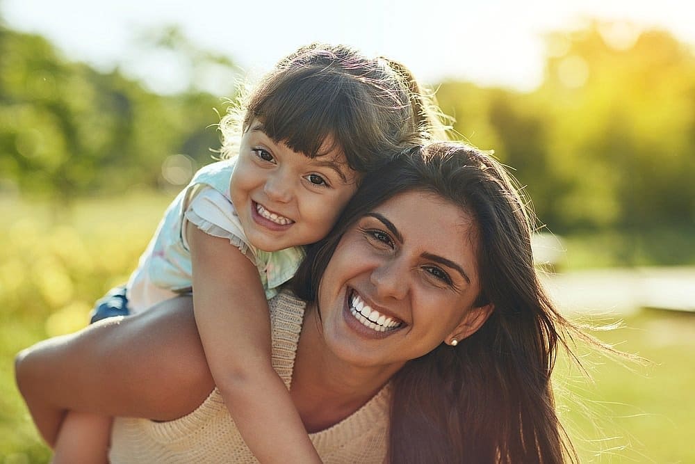 A woman gives a girl a piggyback ride outdoors, both smiling at Carpinello Orthodontics in Newtown Square, PA.