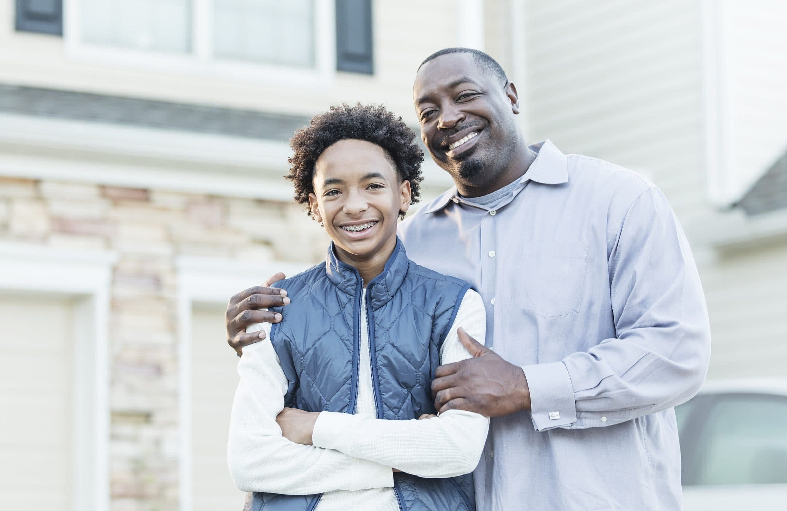 A man and smiling boy stand together outside a suburban house, possibly after a Carpinello Orthodontics consult in Drexel Hill, Newtown Square or Edgmont, PA.