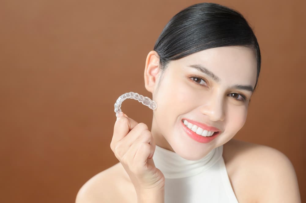 A woman smiles holding an Invisalign aligner, promoting Carpinello Orthodontics in Newtown Square, PA.