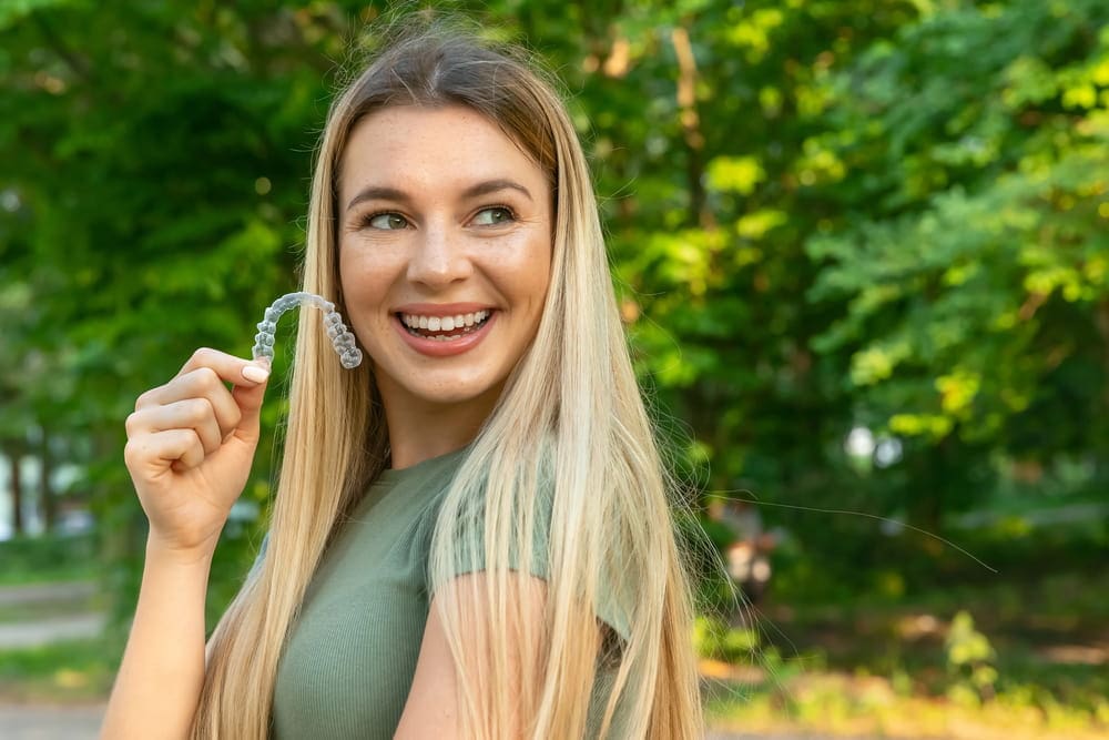Smiling outdoors, a young blonde woman shows an Invisalign from Carpinello Orthodontics in Drexel Hill, PA.