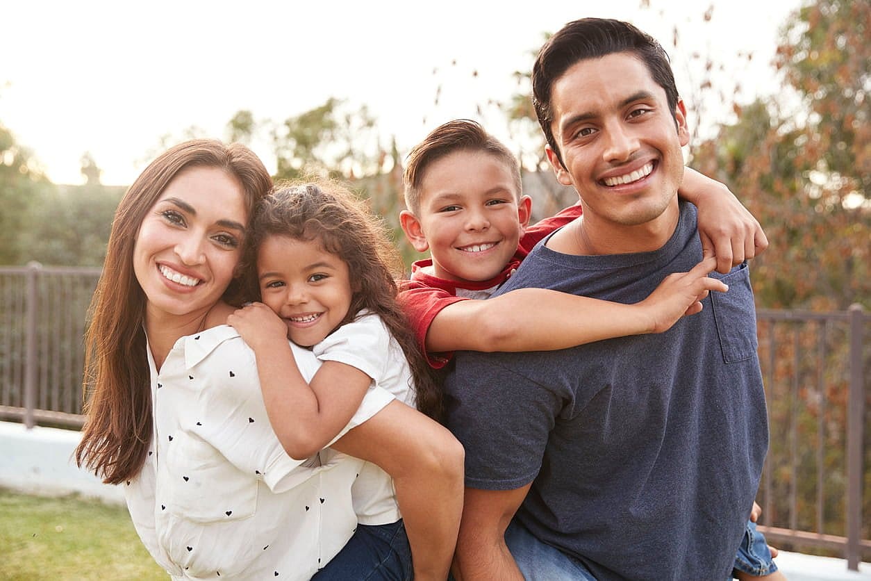 A smiling family of four enjoys a piggyback ride outdoors, kids flashing represent the best Orthodontic Appliances at Carpinello Orthodontics in Newtown Square, PA.