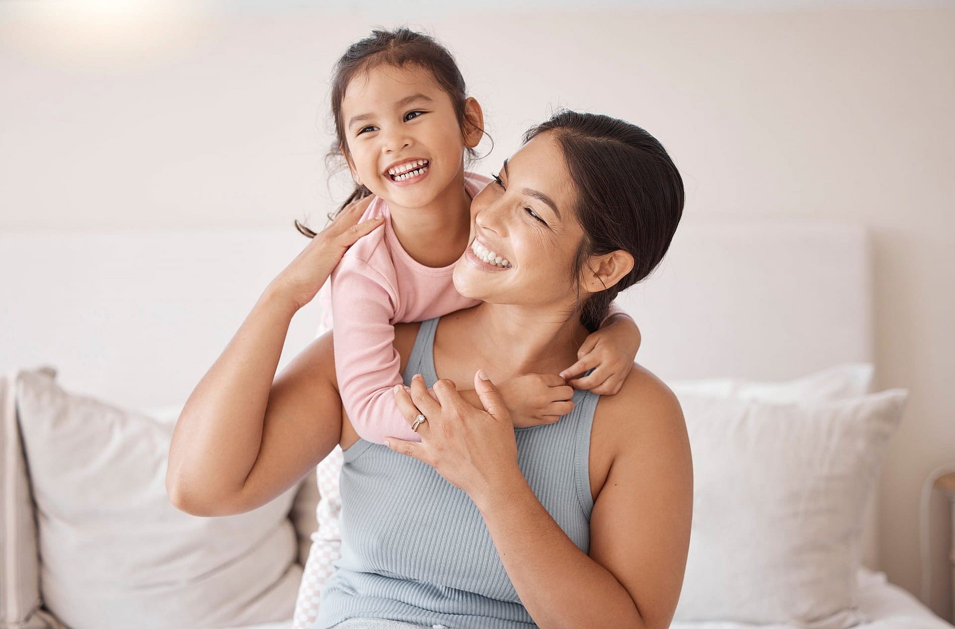 Smiling on a bed, a woman is hugged by a happy girl, representing the best orthodontic appliances from Carpinello Orthodontics in Edgmont, PA.