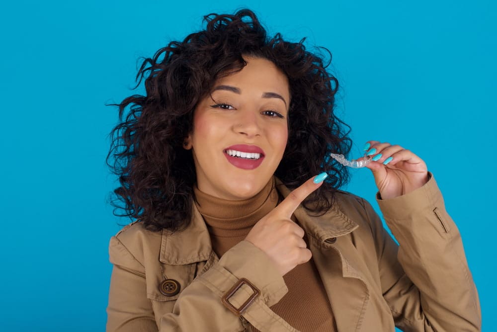 A woman with curly hair smiles and points to an Invisalign aligner, representing Carpinello Orthodontics in Newtown Square, PA.