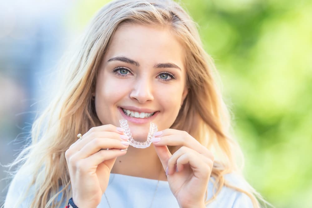 Smiling woman with long blonde hair holds Invisalign aligner outdoors; Carpinello Orthodontics in Drexel Hill, PA.