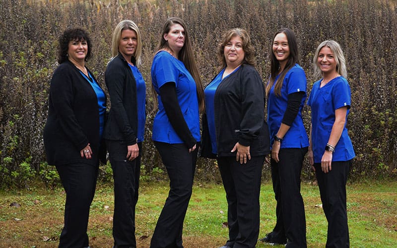 At Carpinello Orthodontics, located in Drexel Hill, Newtown Square or Edgmont, PA, six administrative team women in blue scrubs smile outdoors.