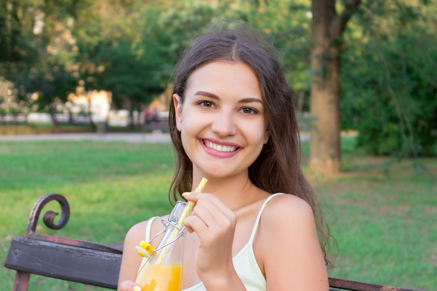 A young woman smiles on a park bench with orange juice, promoting Orthodontists at Carpinello Orthodontics in Drexel Hill, PA.