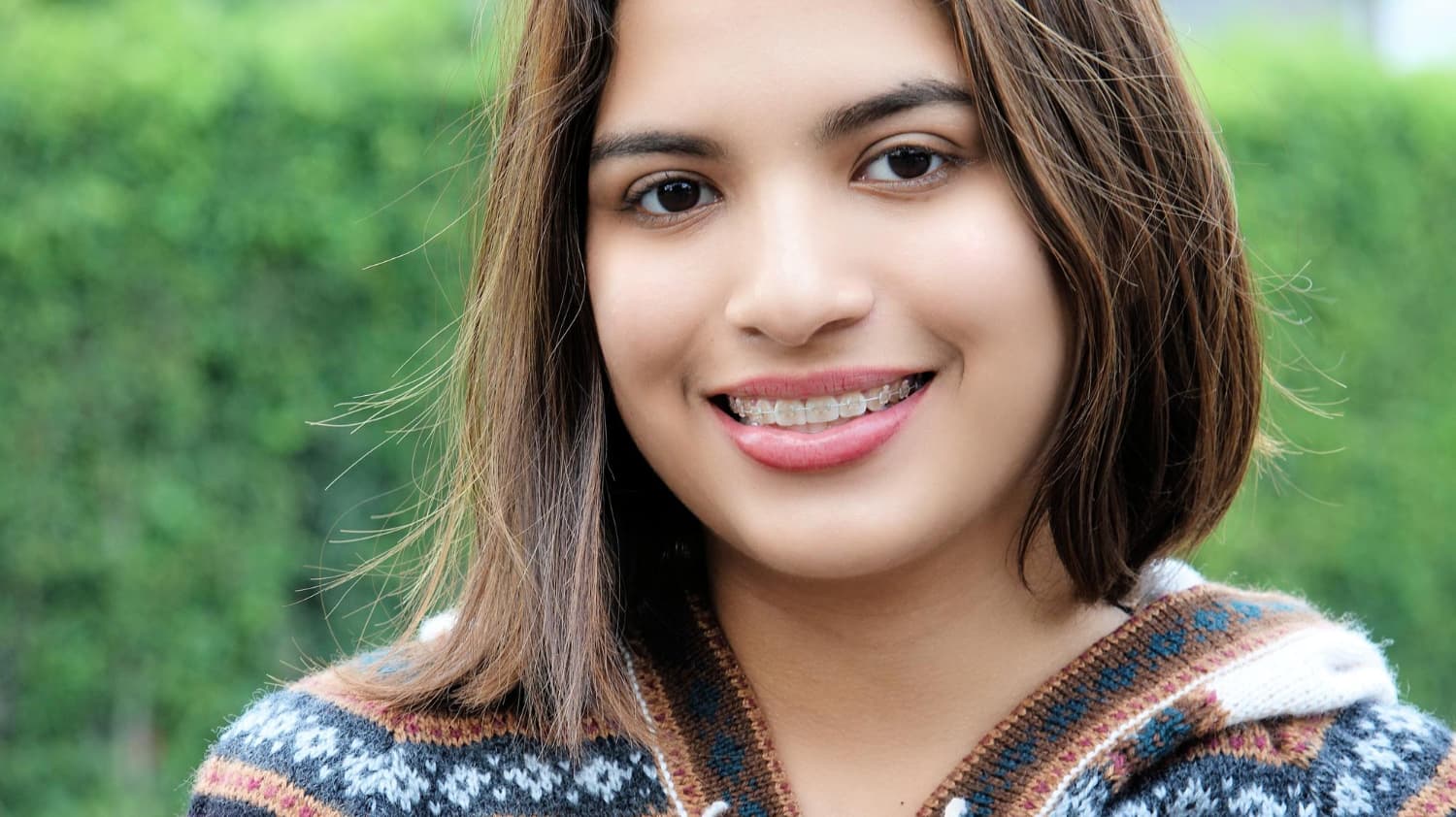 Smiling with clear braces, a brown-haired teen in a patterned sweater is photographed outdoors at Carpinello Orthodontics in Drexel Hill, Newtown Square or Edgmont, PA.