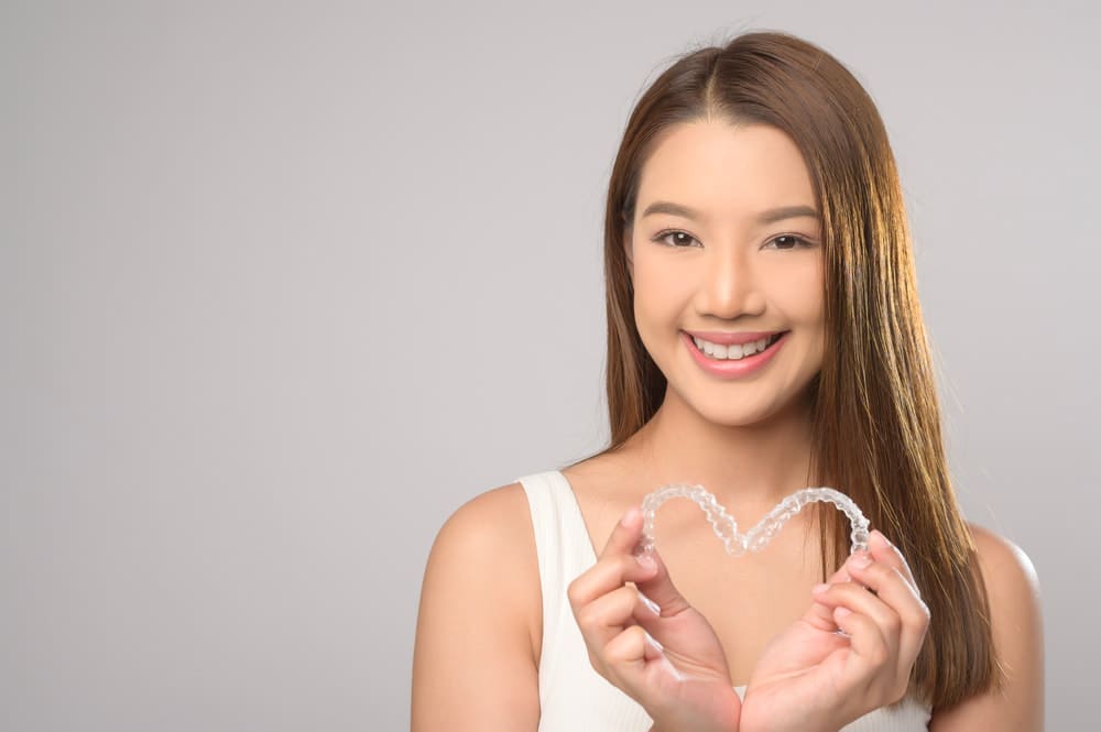 Smiling woman holds heart-shaped Invisalign aligners in front of her chest; Carpinello Orthodontics with flexible payment plans in Drexel Hill, Newtown Square or Edgmont, PA.