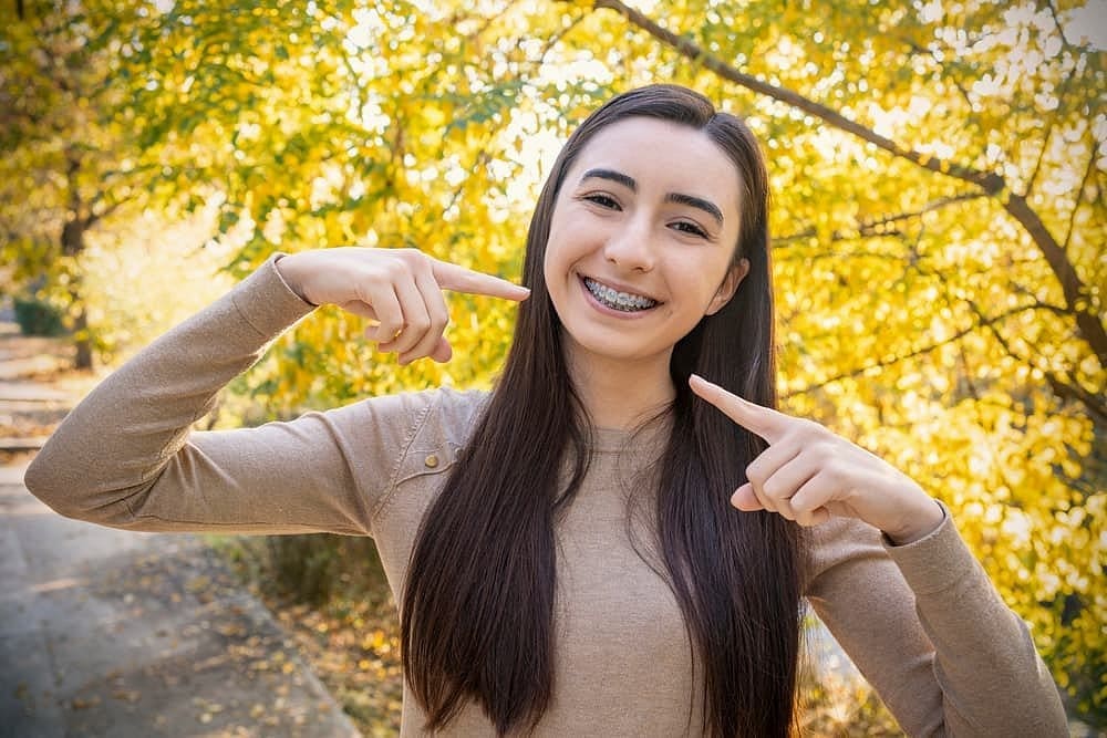 Smiling woman with affordable braces points at her teeth, outdoors near trees. Carpinello Orthodontics in Drexel Hill, Newtown Square or Edgmont, PA.