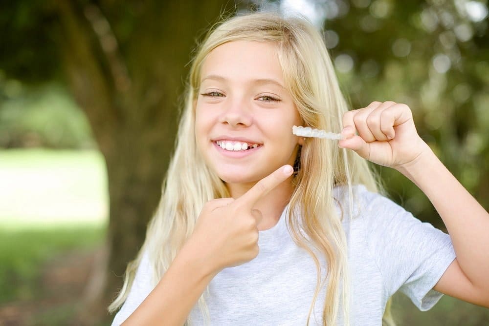 A smiling girl with long blonde hair holds clear aligners, promoting Children’s Orthodontist from Carpinello Orthodontics in Drexel Hill, Newtown Square, or Edgmont, PA.