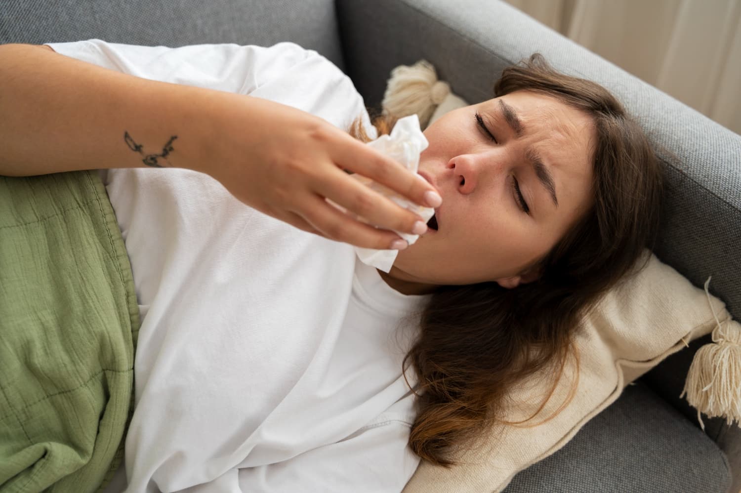 A woman on a couch under a green blanket sneezes into a tissue, showing mouth breathing causes at Carpinello Orthodontics in Drexel Hill, Newtown Square or Edgmont, PA.