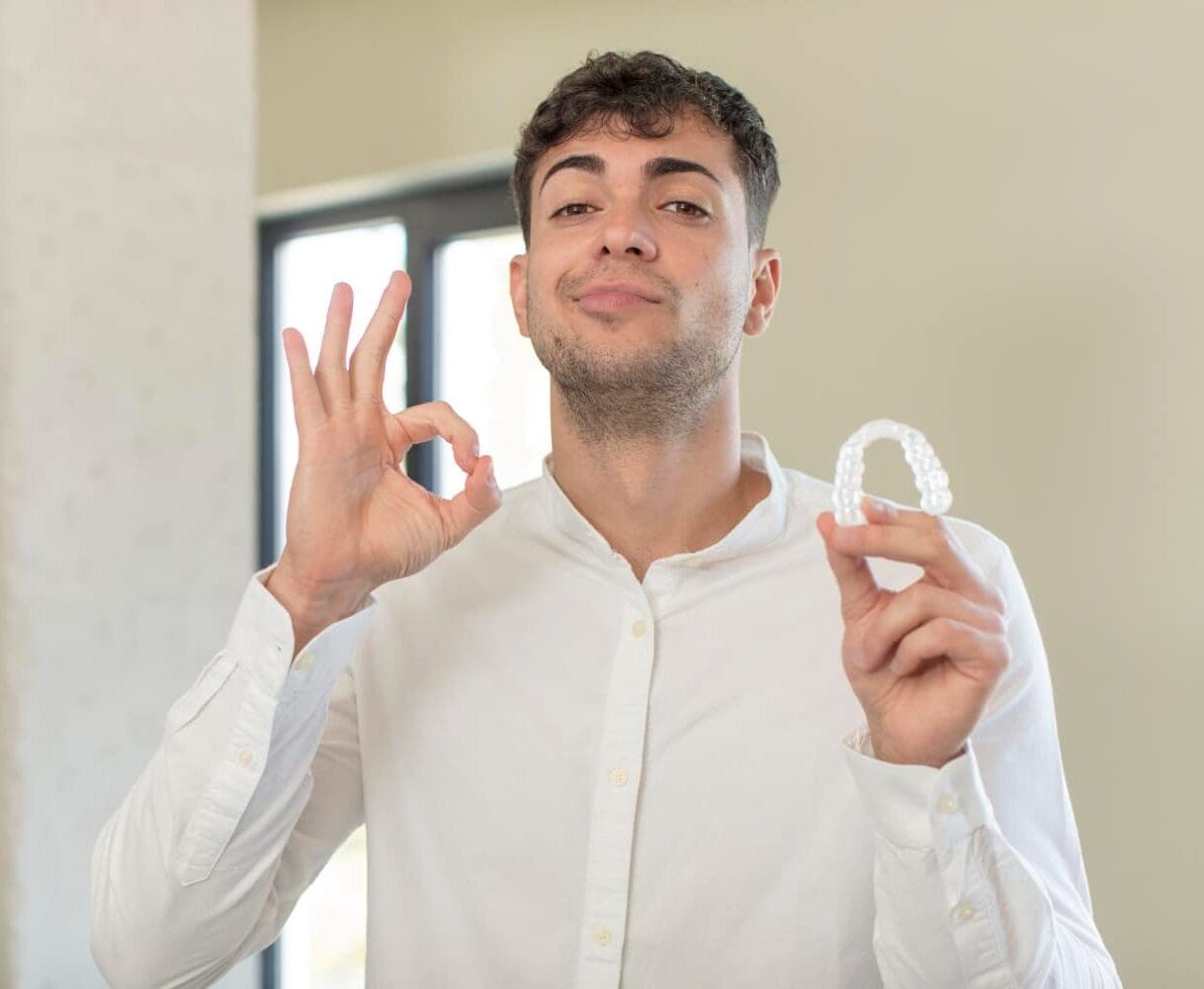 A man indoors holds a clear aligner and makes an OK sign, showcasing Carpinello Orthodontics in Drexel Hill, Newtown Square or Edgmont, PA.