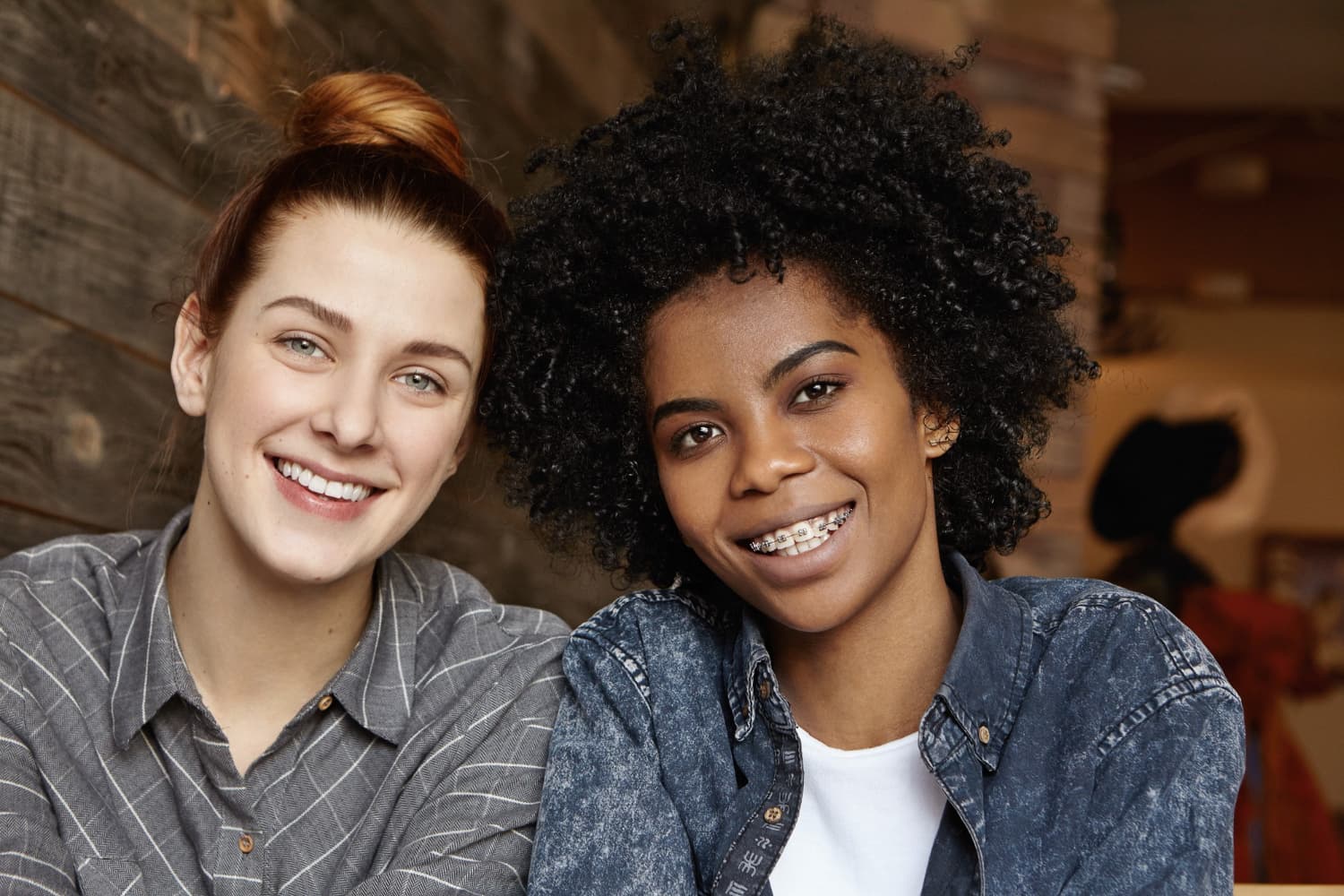 Smiling together, two young women—one with a bun, one with curly hair and affordable braces from Carpinello Orthodontics in Drexel Hill, Newtown Square or Edgmont, PA—pose against a wooden wall.