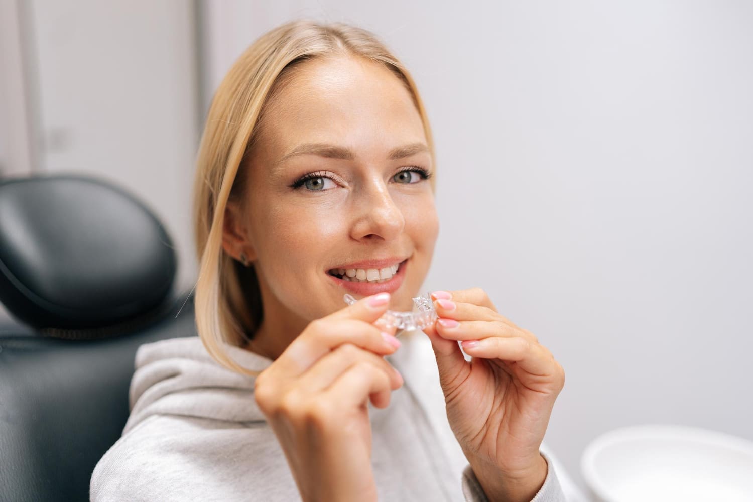A woman in a dental chair smiles, holding a clear aligner, including Invisalign is covered by insurance at Carpinello Orthodontics in Drexel Hill, Newtown Square or Edgmont, PA.