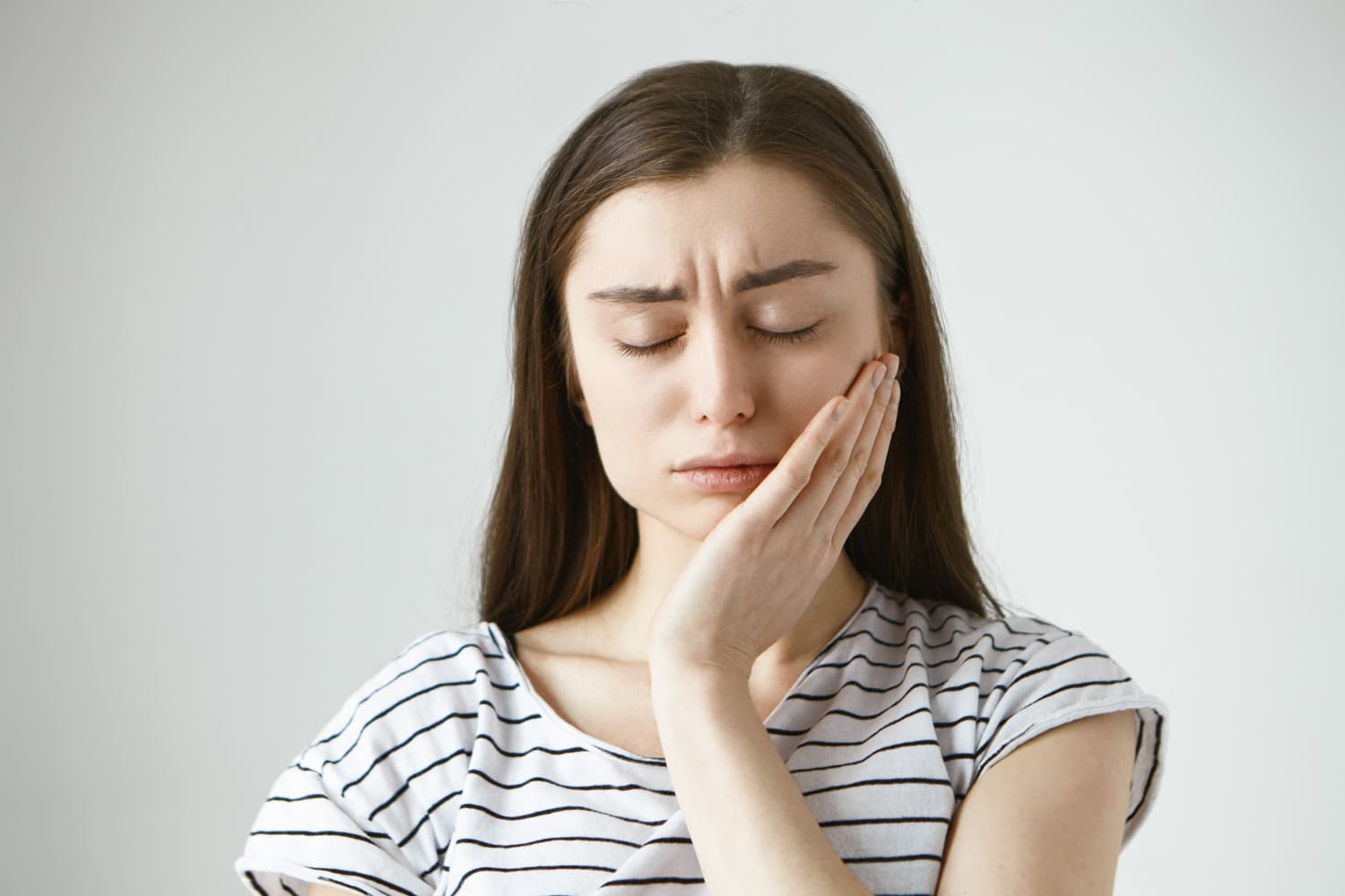 Woman with long brown hair holding her cheek in discomfort, illustrating Invisalign vs braces at Carpinello Orthodontics in Drexel Hill, Newtown Square, or Edgmont PA.