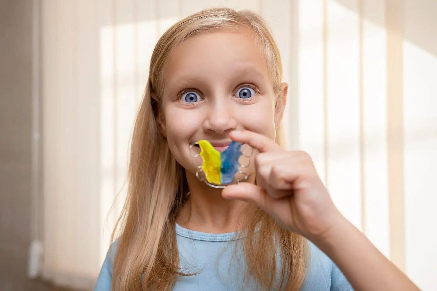 Smiling, a young girl with long blonde hair shows off colorful retainers from a children's orthodontist at Carpinello Orthodontics in Drexel Hill, Newtown Square, or Edgmont, PA.