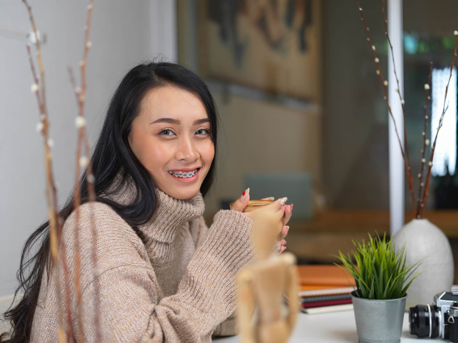 A woman with long black hair and braces, at her desk with plants and a camera, considers Carpinello Orthodontics in Drexel Hill, Newtown Square or Edgmont, PA.