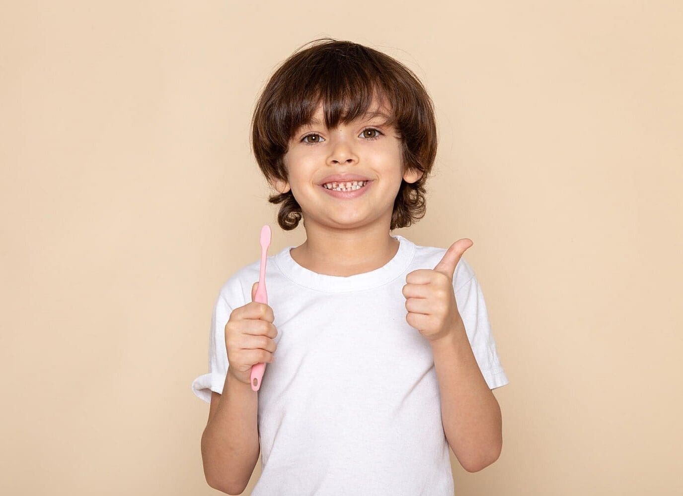 A smiling child in a white shirt holds a pink toothbrush and gives a thumbs up, showing common orthodontic issues in Children and recommending a trusted Children’s Orthodontist from Carpinello Orthodontics in Drexel Hill, Newtown Square, or Edgmont, PA.