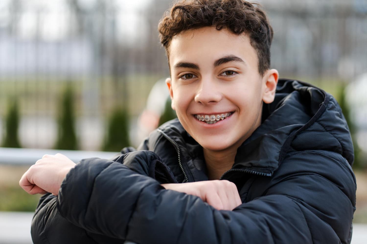 Smiling teen with metal braces at Carpinello Orthodontics in Drexel Hill, Newtown Square or Edgmont, PA, sits outdoors in a black jacket.