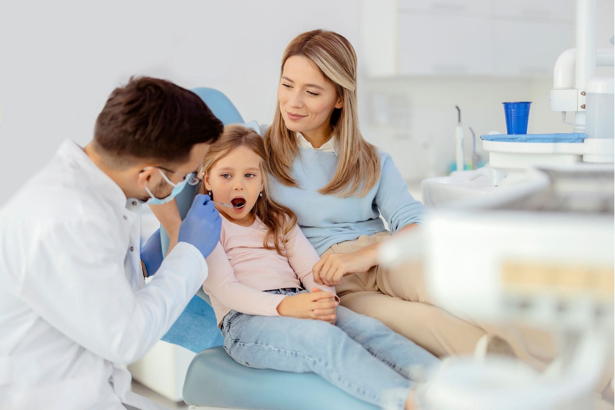 A Children’s Orthodontist at Carpinello Orthodontics in Drexel Hill, Newtown Square, or Edgmont, PA, examines a girl sitting on her mom’s lap to show the Importance of early evaluation checkups with a Children’s Orthodontist.
