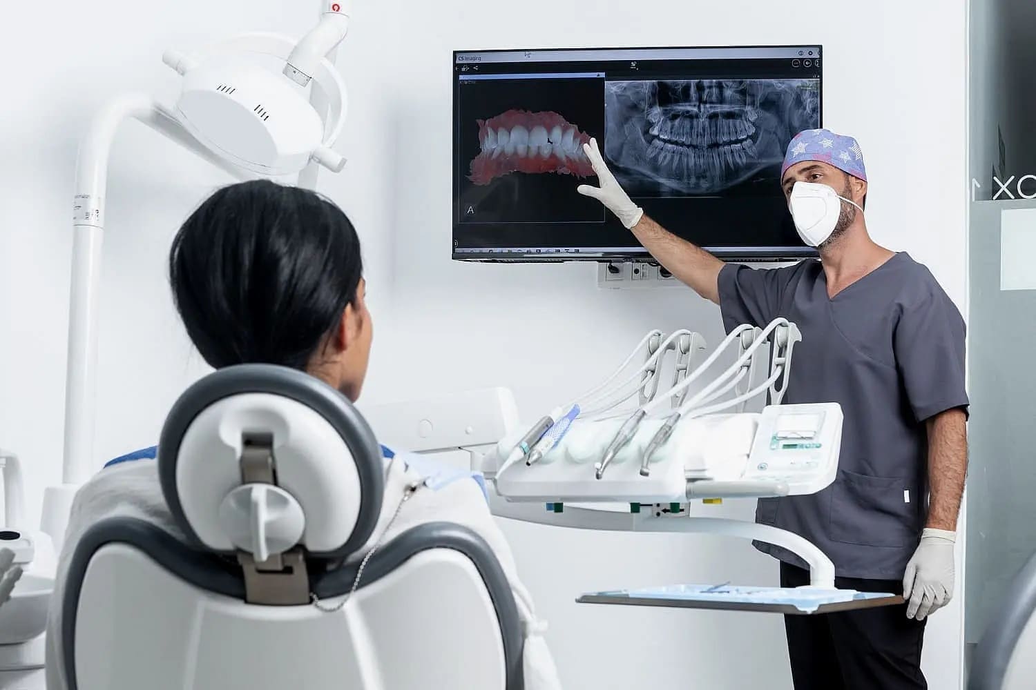 In Carpinello Orthodontics in Drexel Hill, Newtown Square, or Edgmont, PA, a masked dentist shows dental X-rays and future advancements in jaw surgery to a seated patient.