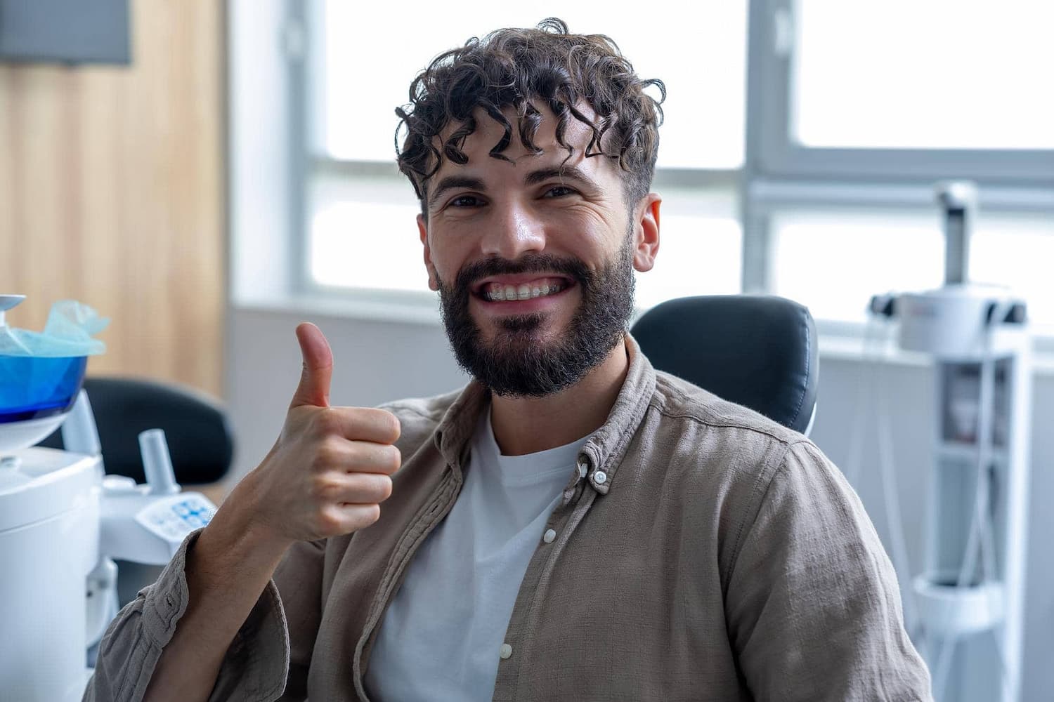 Smiling man with curly hair and beard indoors, showing clear braces from Carpinello Orthodontics in Drexel Hill, Newtown Square or Edgmont, PA, and giving a thumbs up.
