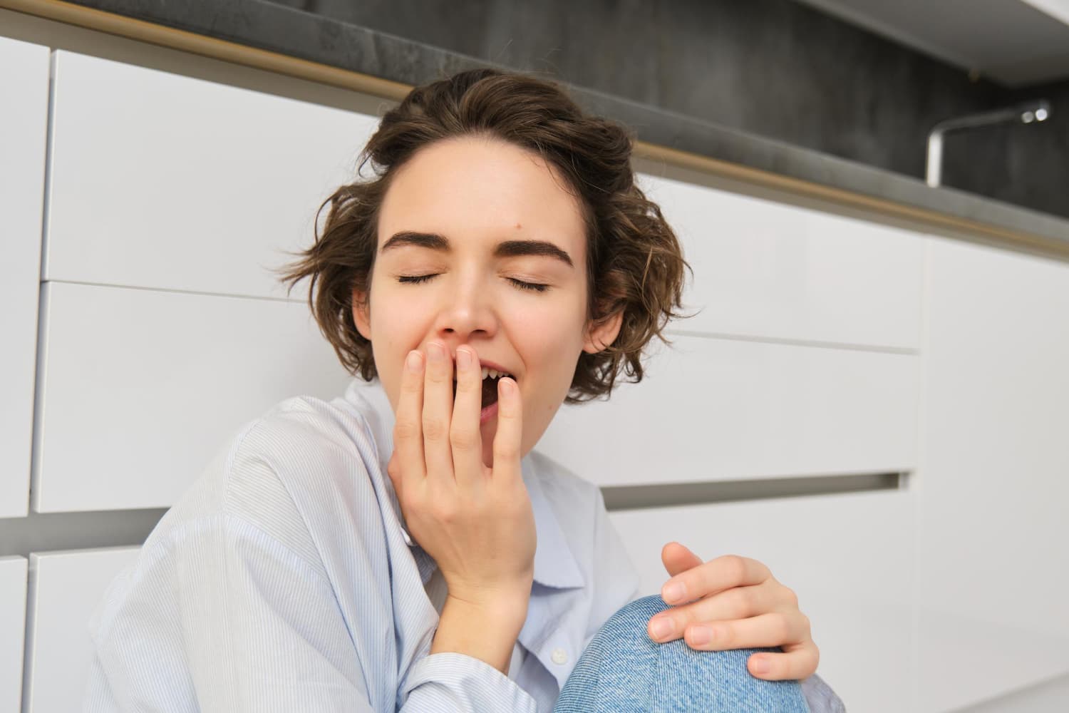 Sitting by white cabinets, a person yawns with hand over mouth—a sign of underbite Carpinello Orthodontics treats in Drexel Hill, Newtown Square or Edgmont, PA.