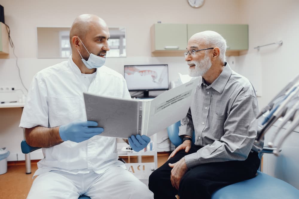 At Carpinello Orthodontics in Drexel Hill, Newtown Square or Edgmont, PA, a dentist reviews a chart and insurance with an older man.