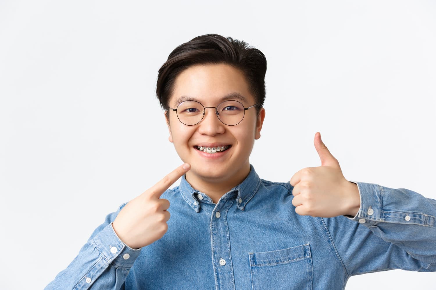 Smiling teen in glasses and denim points to braces, thumbs up, proud of Carpinello Orthodontics' flexible plan in Drexel Hill, Newtown Square or Edgmont, PA.