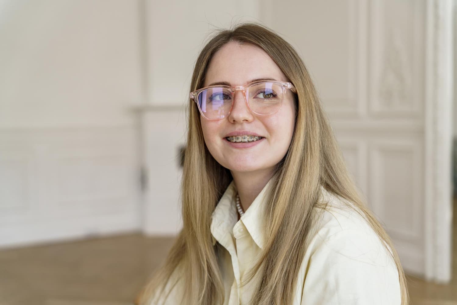 A smiling young woman with long blonde hair, glasses, and adult braces by Carpinello Orthodontics in Drexel Hill, Newtown Square or Edgmont, PA sits indoors against a white wall.