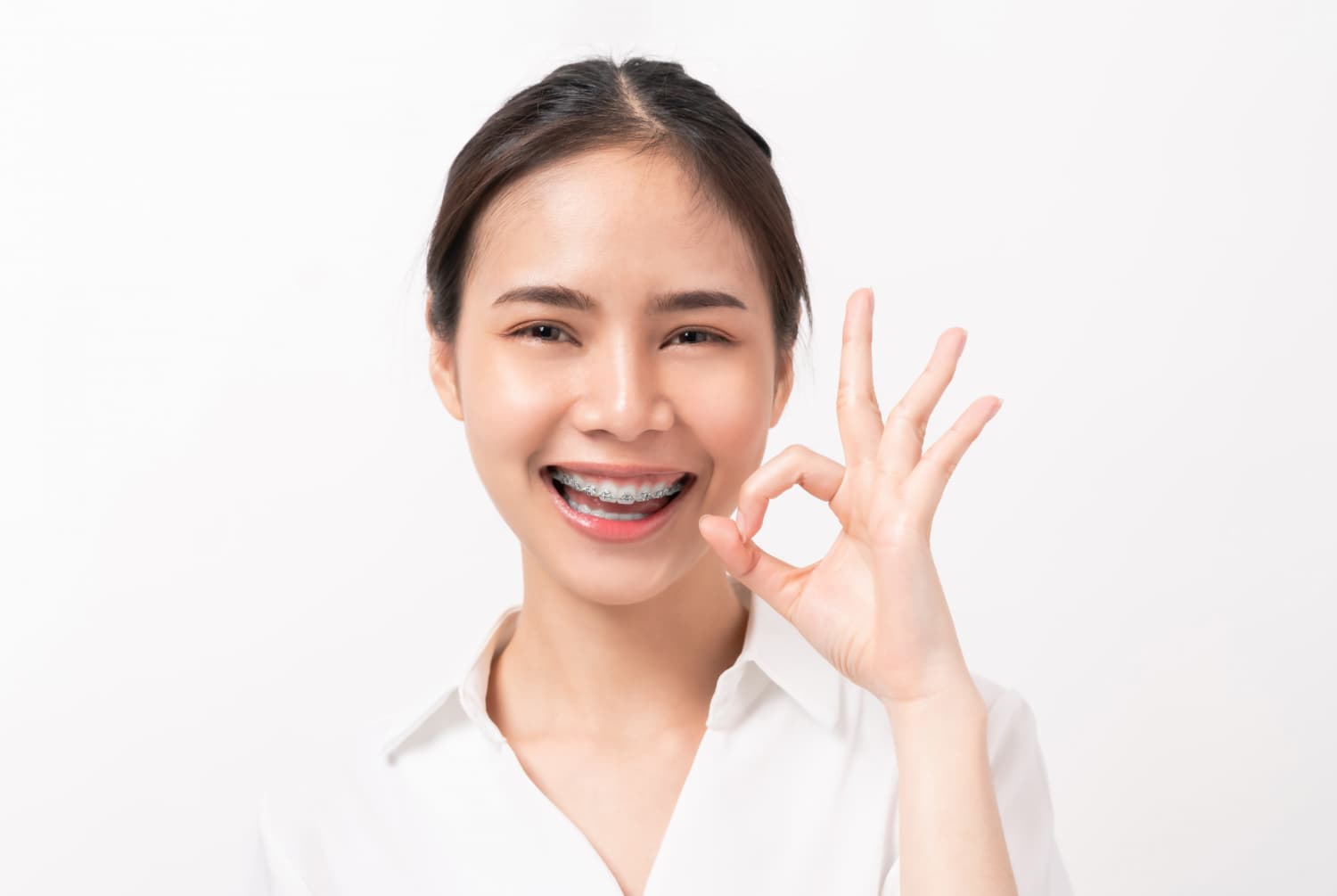 A young woman with braces in a white shirt smiles and makes an OK sign, confident in her Carpinello Orthodontics plan in Drexel Hill, Newtown Square or Edgmont, PA.