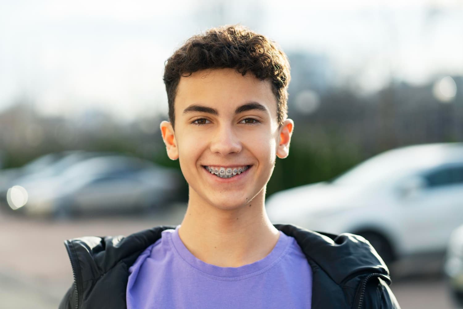 A smiling teen with curly hair and braces outdoors, showing underbite progress at Carpinello Orthodontics in Drexel Hill, Newtown Square or Edgmont, PA.