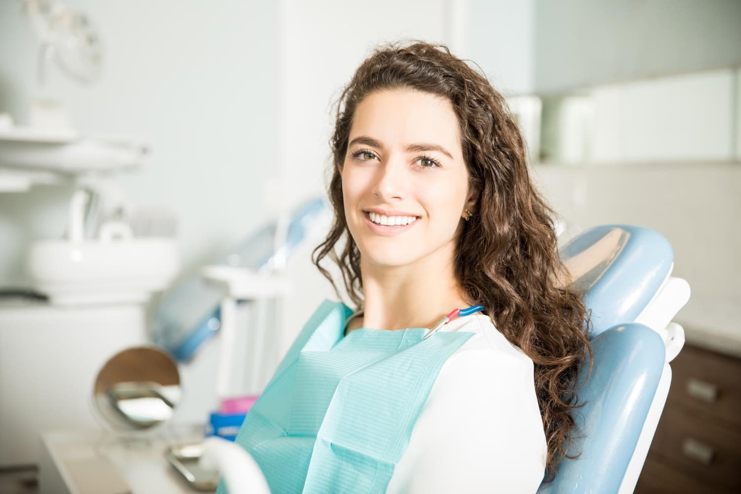 A woman with long curly hair smiles in a dental chair, showing clear braces at Carpinello Orthodontics in Drexel Hill, Newtown Square or Edgmont, PA.