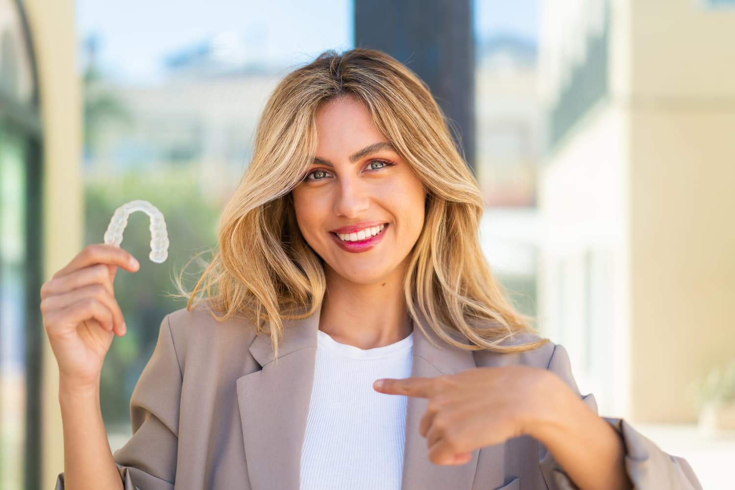 A smiling woman with long blonde hair points to a clear dental aligner, showing adult braces options at Carpinello Orthodontics in Drexel Hill, Newtown Square or Edgmont, PA.