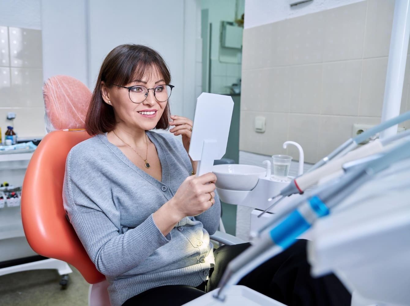 A woman smiles at her reflection in a handheld mirror and finds the right orthodontic treatment as an adult at Carpinello Orthodontics in Drexel Hill, Newtown Square, or Edgmont, PA.
