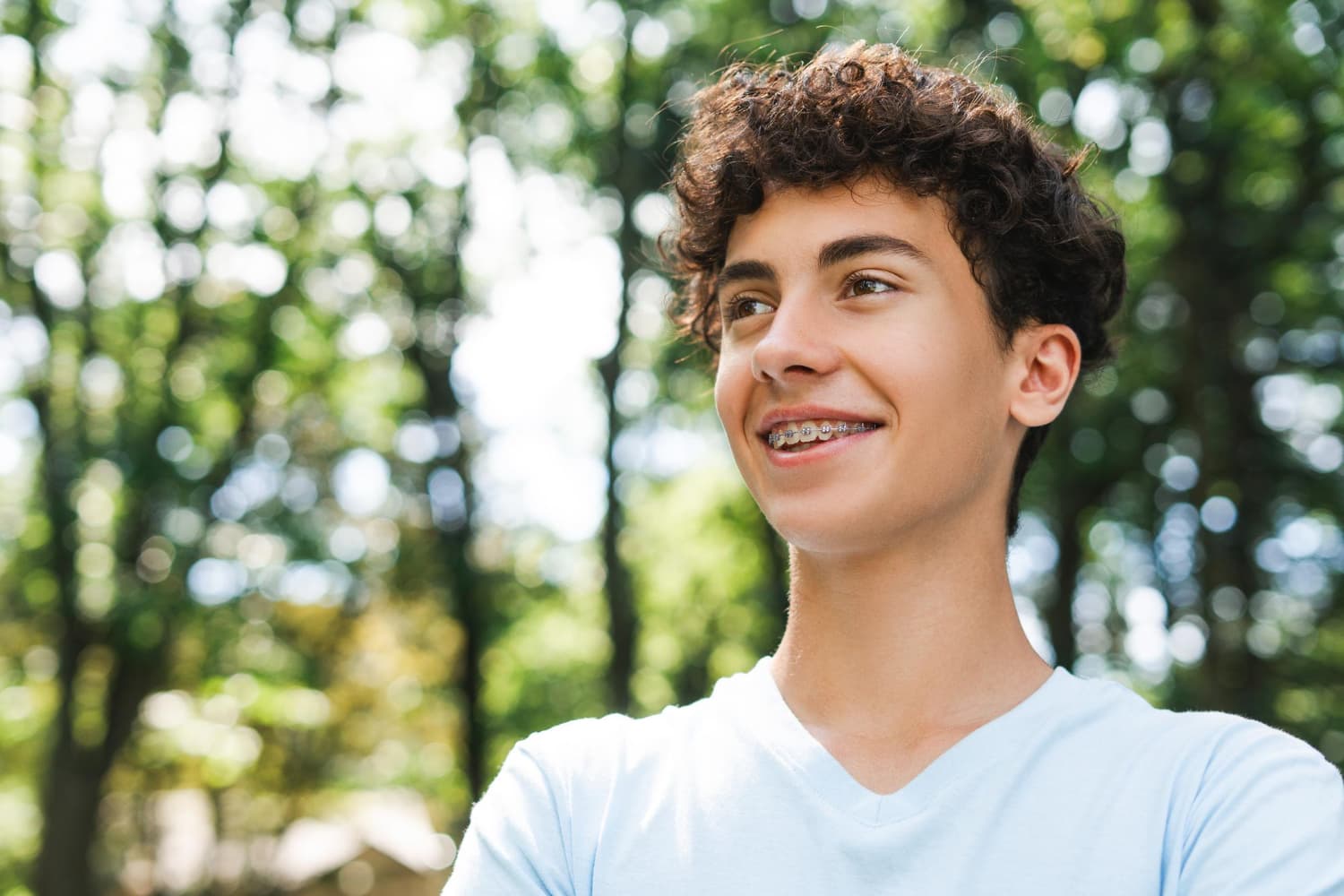 A smiling teen with curly hair and metal braces, outdoors in a light blue T-shirt, represents Carpinello Orthodontics in Drexel Hill, Newtown Square or Edgmont, PA.