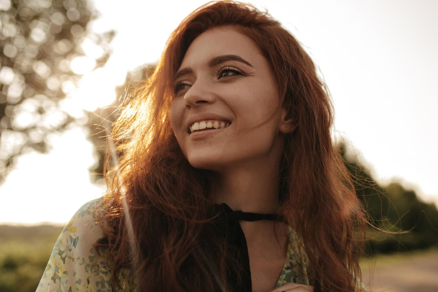 A young red-haired woman smiles confidently outdoors, sunlight on her face, after affordable braces at Carpinello Orthodontics in Drexel Hill, Newtown Square or Edgmont, PA.
