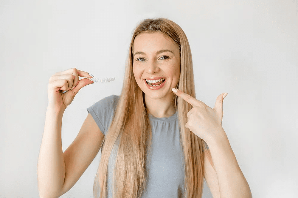 Smiling woman with long blonde hair points to her teeth and holds a clear aligner at Carpinello Orthodontics in Drexel Hill, Newtown Square or Edgmont, PA, promoting Invisalign cost benefits.