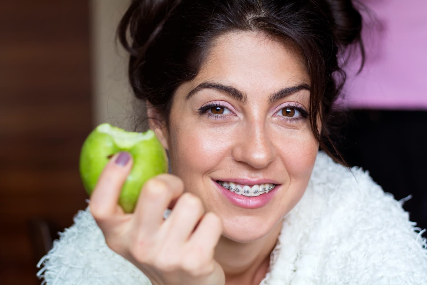 A woman with braces smiles, holding a bitten green apple, promoting overbite correction at Carpinello Orthodontics in Drexel Hill, Newtown Square or Edgmont, PA.