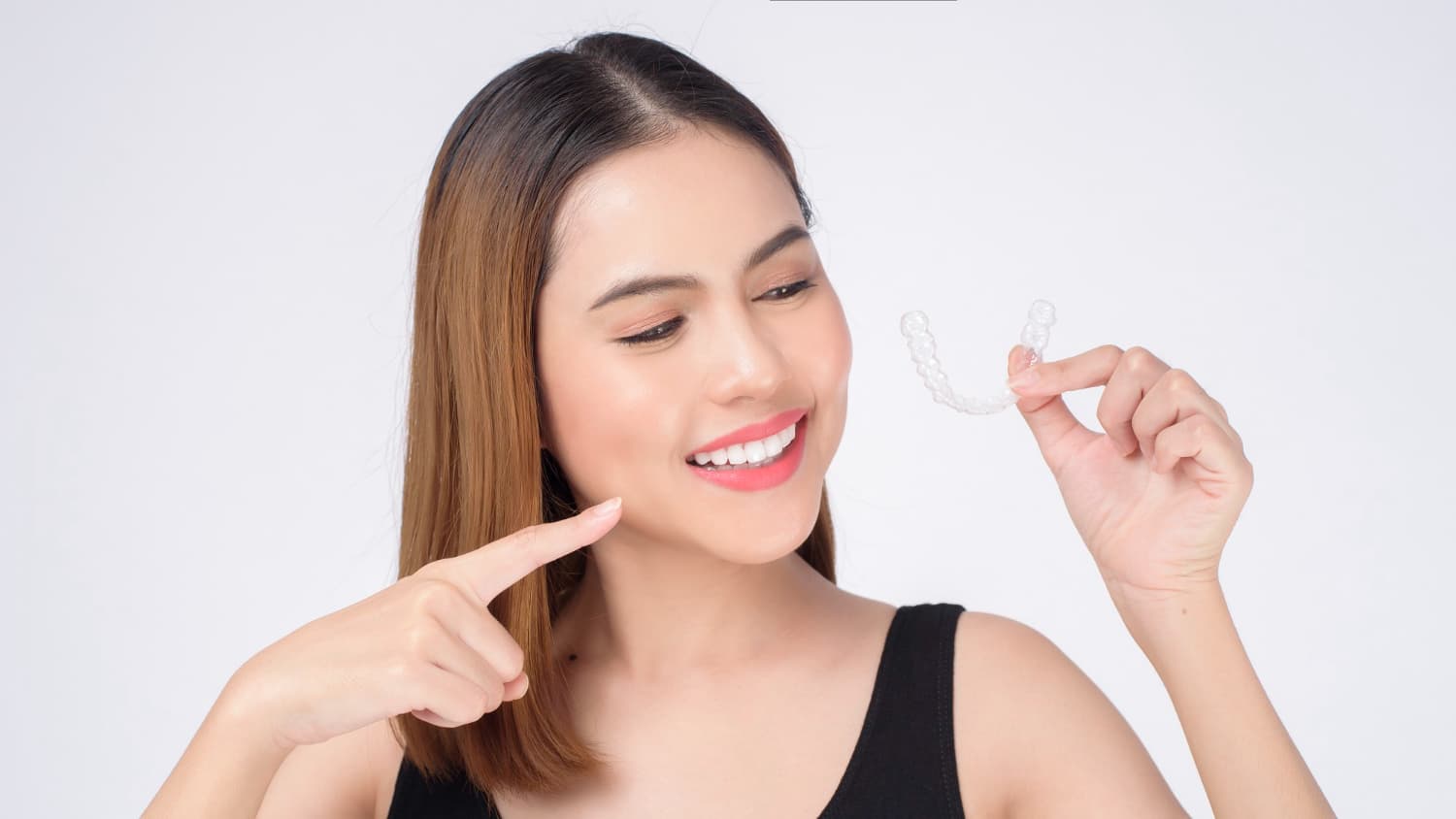 Smiling woman pointing at her teeth while holding a clear aligner, highlighting that Carpinello Orthodontics offers adult braces alternatives and accepts dental insurance in Drexel Hill, Newtown Square, or Edgmont PA.