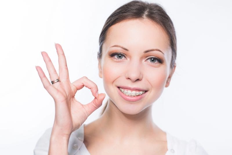 Smiling woman with adult braces gives "OK" gesture in white shirt; Carpinello Orthodontics in Drexel Hill, Newtown Square or Edgmont, PA.