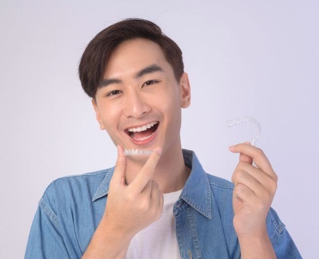 A young man holds a clear aligner and points to his teeth, highlighting affordable Invisalign at Carpinello Orthodontics in Drexel Hill, Newtown Square or Edgmont, PA.