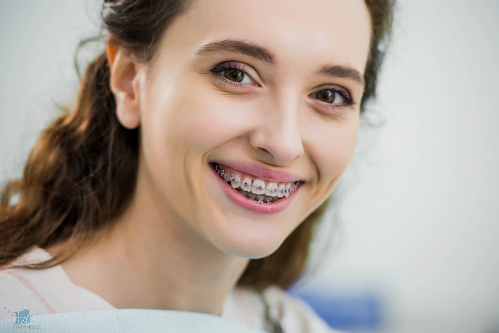 A smiling girl holds and points to an Invisalign Teen aligner with Carpinello Orthodontics in Drexel Hill, Newtown Square or Edgmont, PA, on a blue background.
